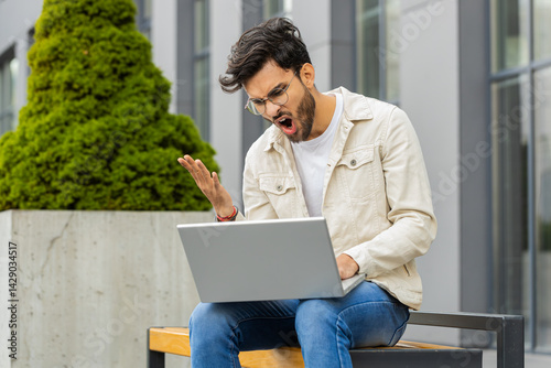 Displeased Indian man use laptop typing browsing working, loses becoming surprised sudden lottery results bad news fortune loss game fail computer virus. Arabian young guy sitting on street city bench