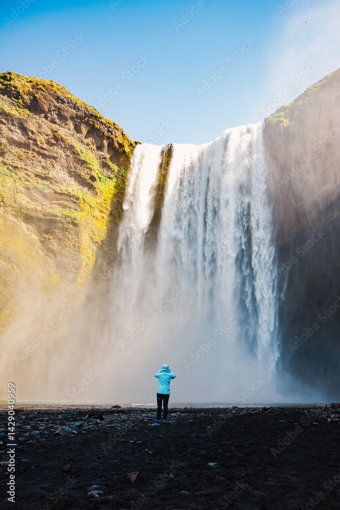 Traveler in blue jacket admiring Skógafoss waterfall in Iceland, surrounded by volcanic rocks and powerful water spray.