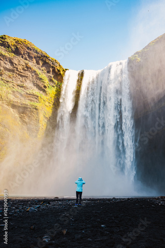 Obraz na płótnie Traveler in blue jacket admiring Skógafoss waterfall in Iceland, surrounded by volcanic rocks and powerful water spray. w salonie