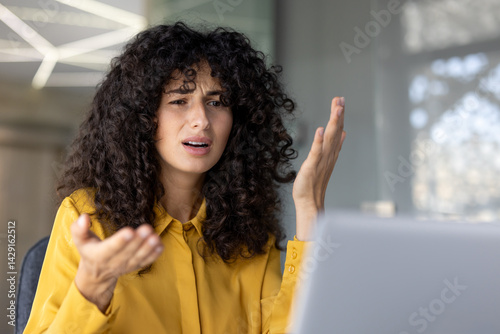 A frustrated woman with curly hair gestures emphatically during a video call, looking upset and confused. photo