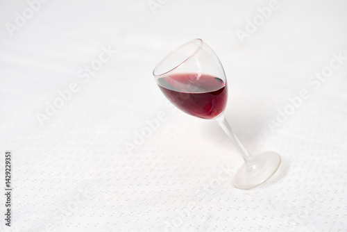 Wine glass with red liquid rests tipped on white tablecloth. Subtle lighting enhances contrast between dark wine and bright background, creating minimalist, elegant scene