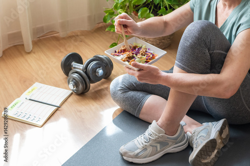 Sitting on a yoga mat, a mature woman enjoys a healthy salad after her home workout, demonstrating a commitment to fitness and balanced nutrition photo
