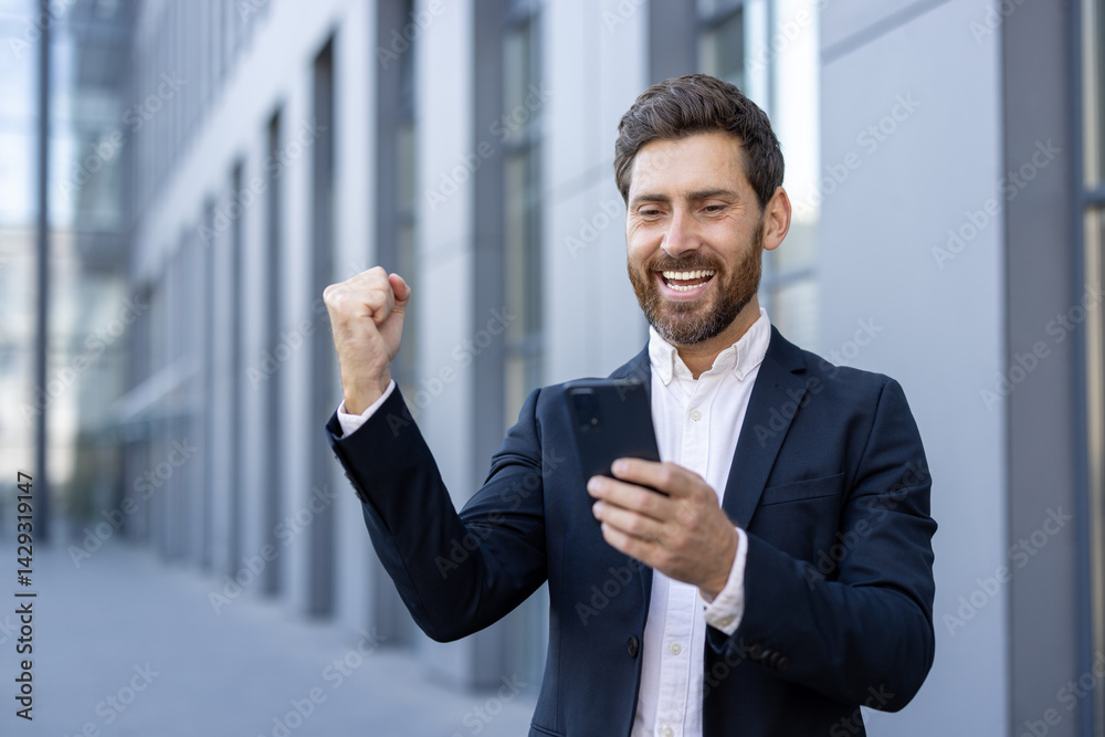 A successful businessman celebrates a win, looking at his phone, with a raised fist and a joyful expression near a modern building.