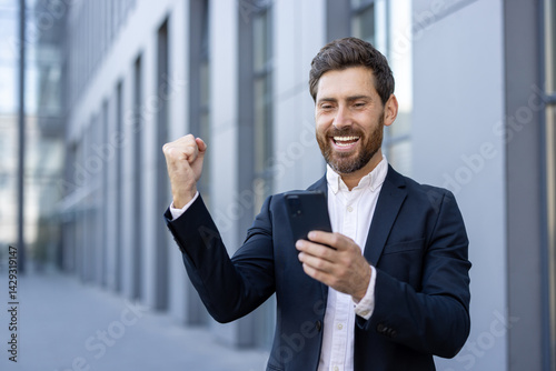 Obraz na płótnie A successful businessman celebrates a win, looking at his phone, with a raised fist and a joyful expression near a modern building. w salonie