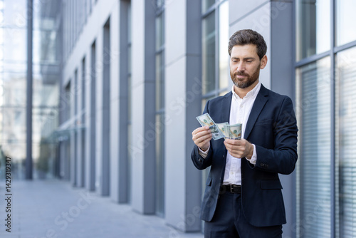 Obraz na płótnie A businessman in a suit counts money in front of a modern building, highlighting wealth and financial success. The man appears thoughtful as he handles the cash. w salonie