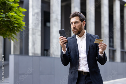 A businessman looks shocked while holding a credit card and his phone, possibly due to a financial issue.