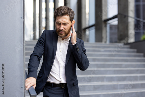 A concerned businessman in a dark suit touches his ear, standing near a modern building's staircase, showing visible stress.