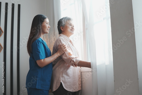Obraz na płótnie An elderly Asian woman stands at her bedroom window looking and thinking about things and an Asian nurse is taking care of her in a nursing home in the morning. w salonie