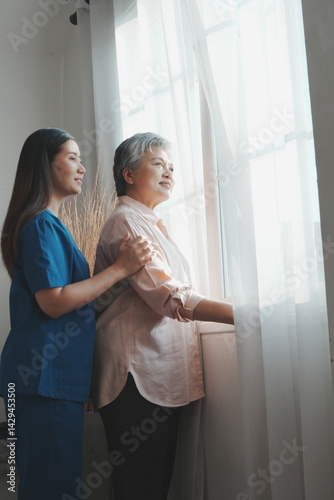 An elderly Asian woman stands at her bedroom window looking and thinking about things and an Asian nurse is taking care of her in a nursing home in the morning.