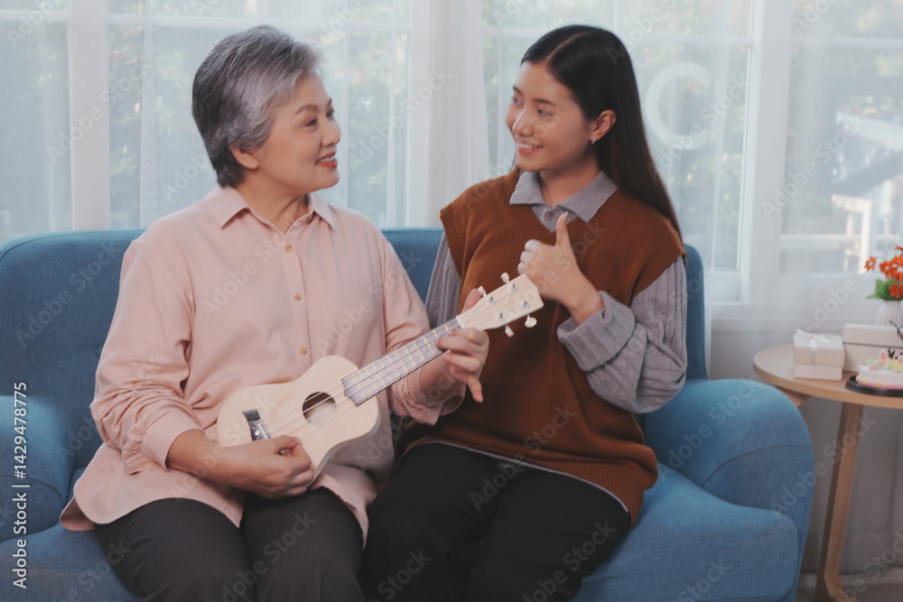 Asian elderly mother and her beautiful 29-year-old daughter relax on the sofa playing ukulele happily together at home.