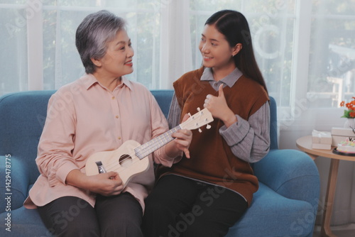 Obraz na płótnie Asian elderly mother and her beautiful 29-year-old daughter relax on the sofa playing ukulele happily together at home. w salonie