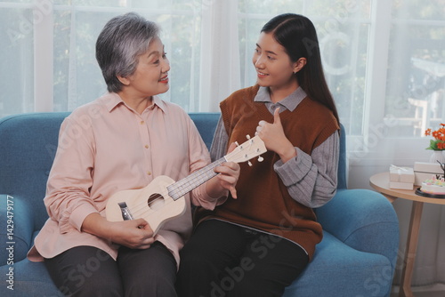 Obraz na płótnie Asian elderly mother and her beautiful 29-year-old daughter relax on the sofa playing ukulele happily together at home. w salonie