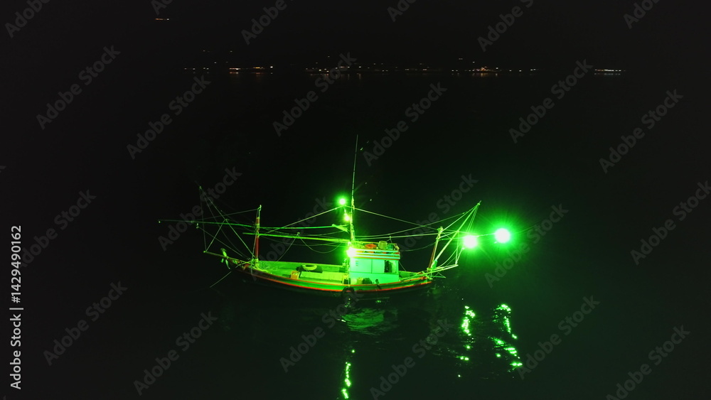 Aerial view of a fishing boat using green lights for night fishing, creating a mesmerizing scene with the reflection on the water and the distant lights of Koh Phangan island in the background