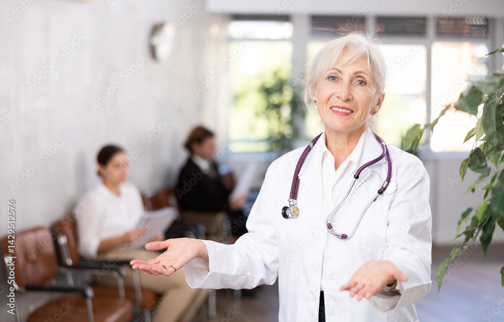 Fototapeta premium Smiling friendly elderly female doctor in white coat making welcome gesture, politely inviting patients in medical office ..