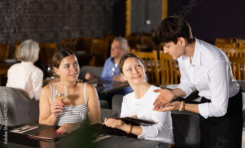 Two interested positive female guests sitting at table in restaurant, holding menu and talking to polite young waiter about dishes choice