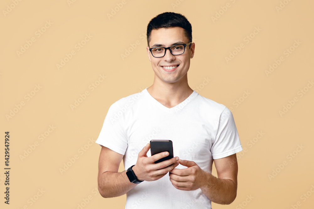 Man look at smartphone, feel joyful reading good news. Smiling young blogger student in glasses, white t-shirt with smart watch search information and looking at camera, isolated on light background Man look at smartphone, feel joyful reading good news. Smiling young blogger student in glasses, white t-shirt with smart watch search information and looking at camera, isolated on light background
