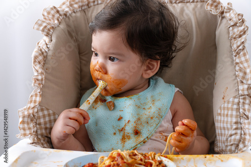 Adorable messy little child girl hungry use hand eating spaghetti sitting in high-powered chair at home. Toddler child dirty with tomato sauce making mess face looking at food. Self-feeding conceptper