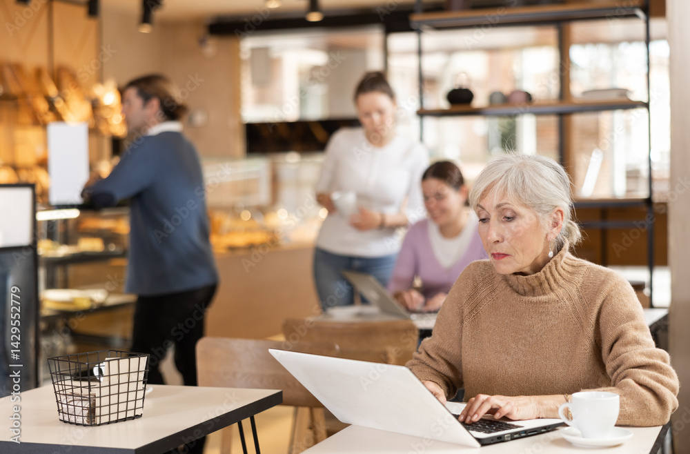 Elderly woman sitting at table, using laptop and enjoing cup of tea in coffee house Elderly woman sitting at table, using laptop and enjoing cup of tea in coffee house