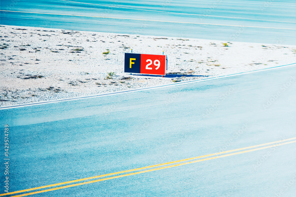 Runway marker sign stands beside with yellow lines, casting a shadow on the gravel edge. Light-blue runway surface extends into the distance, indicating airport area