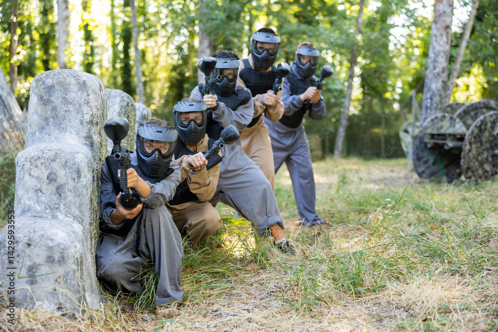 Fototapeta premium Group of men and women in protective clothes and helmets shooting with paintball markers while hiding behind cover. Teams fighting on battlefield outdoors.