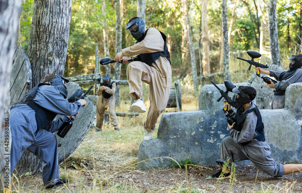 Fototapeta premium Skillful paintball team in protective outwear and helmets attacking hostile positions, running and jumping over enemy covers.