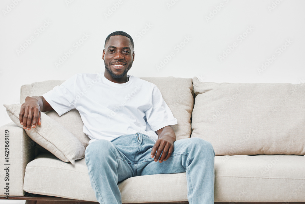 Smiling young Black man relaxing on a beige couch, wearing a casual white t shirt and light blue jeans, exuding a feeling of comfort and happiness Smiling young Black man relaxing on a beige couch, wearing a casual white t shirt and light blue jeans, exuding a feeling of comfort and happiness