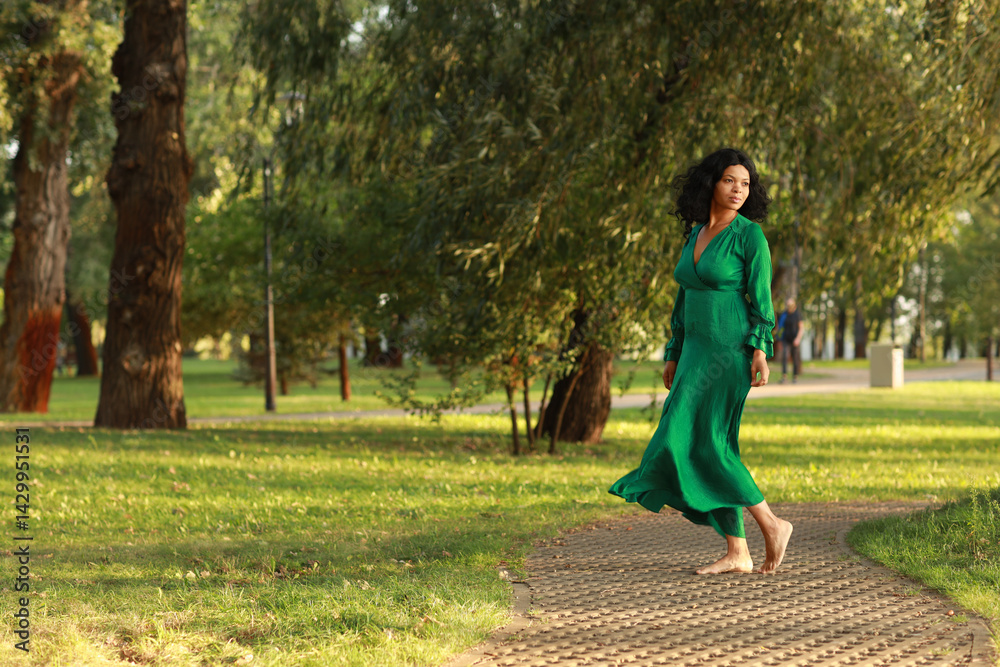Fototapeta premium Young barefoot curly dark haired woman in green dress walking in the park
