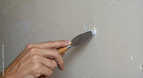 Hand applying putty to wall crack with metal spatula. Close-up of repair technique on white surface. Home maintenance and drywall restoration solutions