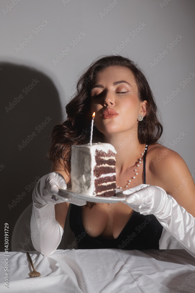 A glamorous woman in an evening dress and white gloves blows out a candle on a slice of birthday cake. This high-quality stock photo is perfect for celebrating special occasions, elegance, and luxury