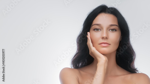 A poised woman with long black hair gently touches her face, showcasing flawless skin against a neutral background. Ideal for beauty, skincare, and cosmetic advertisements, emphasizing natural