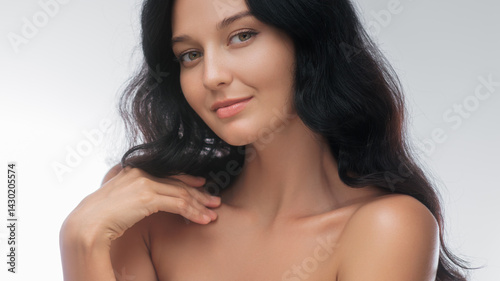 Obraz na płótnie Elegant brunette woman with bare shoulders poses gracefully in a studio setting. Her soft smile and relaxed expression highlight her natural beauty, making this image ideal for beauty, skincare, and w salonie