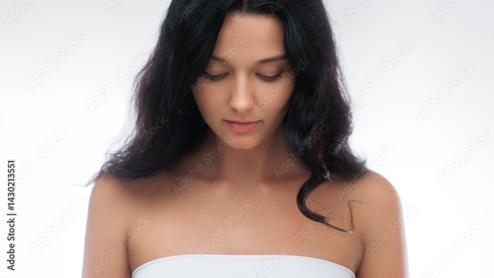 Tranquil brunette woman wearing a white top is captured looking down in a serene studio portrait. The minimalist backdrop and soft lighting enhance her natural beauty, making this image ideal for