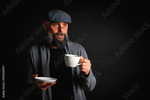 Obraz na płótnie Stylish bearded man in cap and glasses holding coffee cup and saucer on dark background. Thoughtful expression, cozy casual wear, modern vintage atmosphere, morning tea drinking w salonie