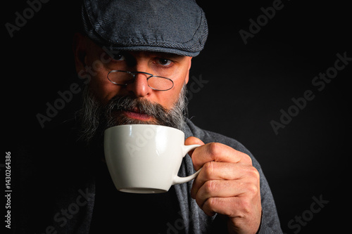 Obraz na płótnie Stylish bearded man in cap and glasses holding coffee cup and saucer on dark background. Thoughtful expression, cozy casual wear, modern vintage atmosphere, morning tea drinking w salonie