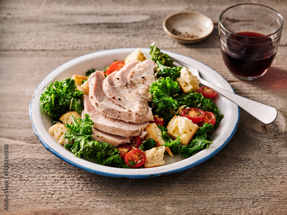 A vibrant turkey kale salad topped with sliced turkey breast, cherry tomatoes, and croutons is presented on a rustic plate. A glass of red beverage adds to the inviting atmosphere A vibrant turkey kale salad topped with sliced turkey breast, cherry tomatoes, and croutons is presented on a rustic plate. A glass of red beverage adds to the inviting atmosphere