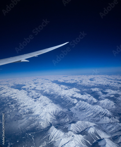 aerial view from an airplane window of the snow covered rocky mountains of western canada mountain range with airplane wing and blue sky vertical air travel adventure background image room for type