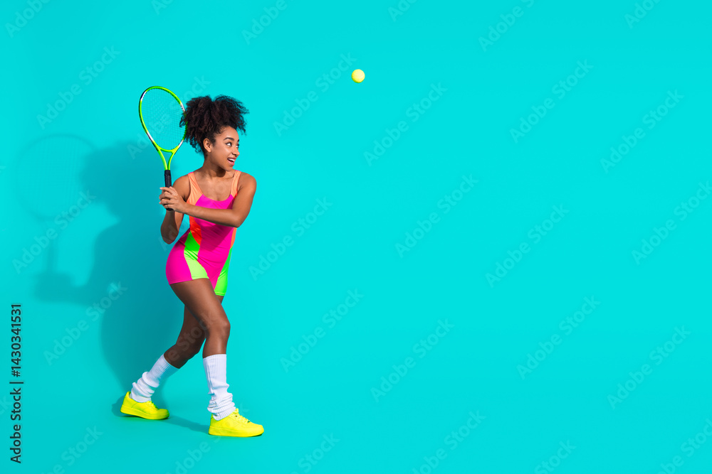 Fototapeta premium Young athletic woman in colorful activewear posing with a tennis racket on a bright teal background promoting fitness