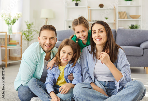 Portrait of happy smiling young family of four with two kids boy and girl sitting on floor at home and looking cheerful at camera. Parents with children enjoying weekend. Family and leisure concept.