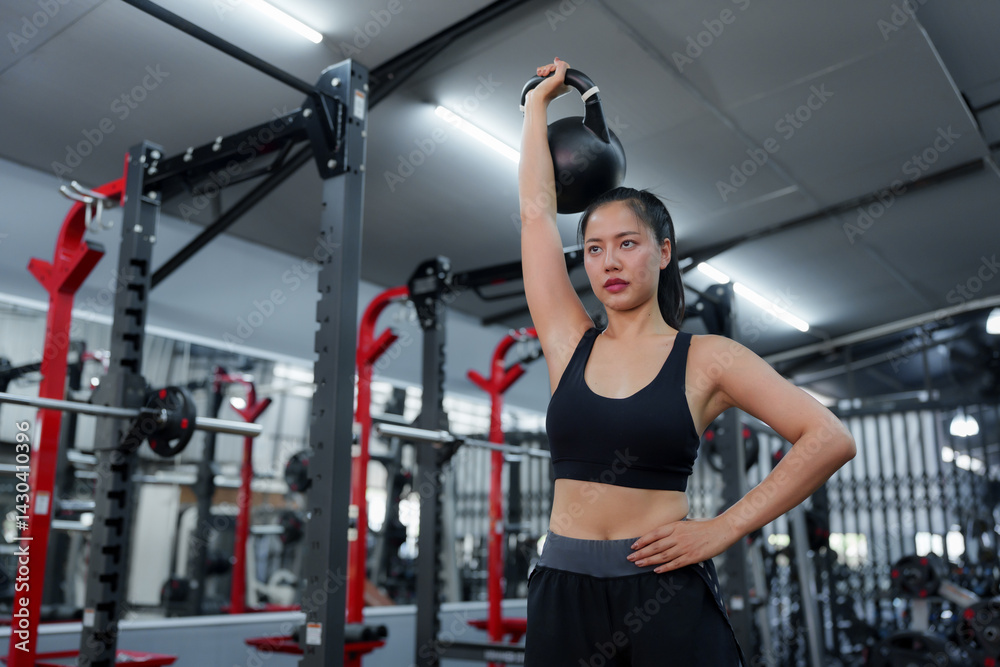 Fototapeta premium Young sportswoman is lifting a kettlebell over her head in a brightly lit modern gym, demonstrating strength and dedication to her fitness routine