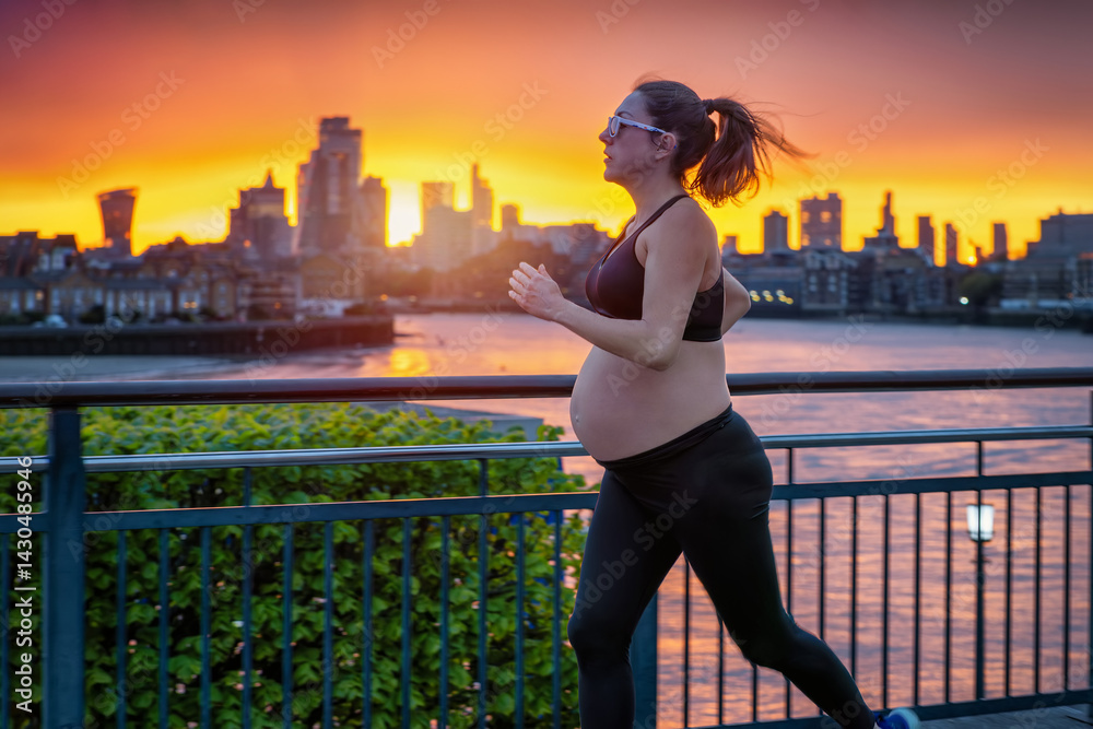 A pregnant woman in sports clothing running for exercise in front of an urban skyline during a fiery sunset