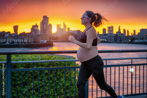 Obraz na płótnie A pregnant woman in sports clothing running for exercise in front of an urban skyline during a fiery sunset w salonie