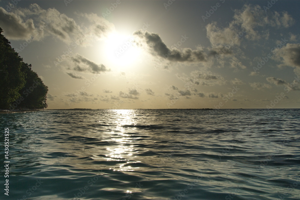 Obraz premium Serene ocean sunset with silhouetted trees and tranquil waves under cloudy sky in the Maldives 