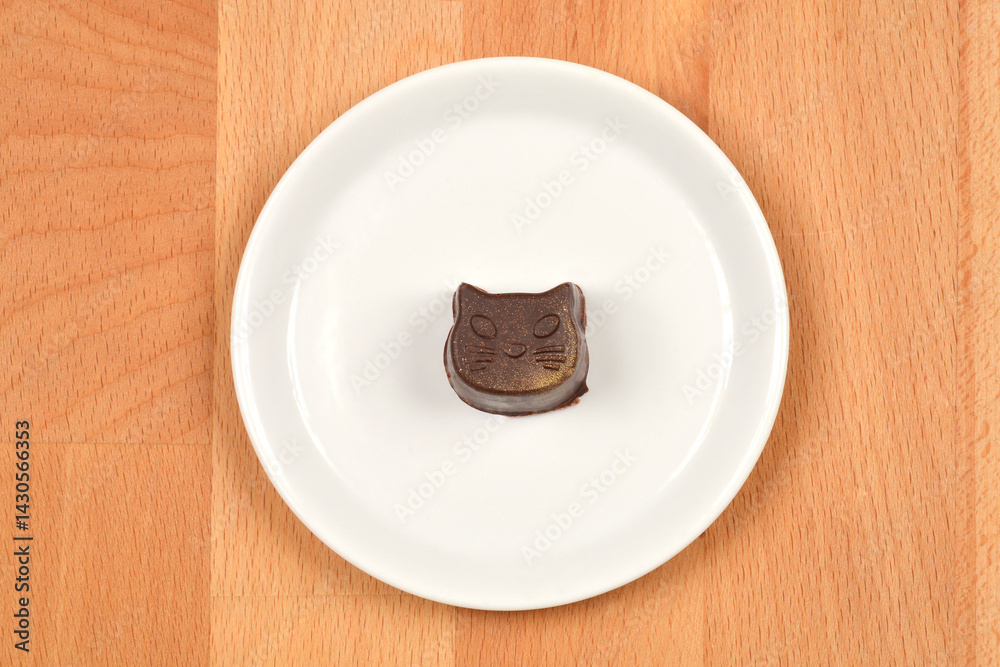 A single, dark homemade praline shaped like a cats face sits on a small white plate against a light wooden background