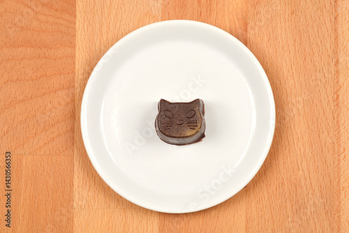 Obraz na płótnie A single, dark homemade praline shaped like a cats face sits on a small white plate against a light wooden background w salonie