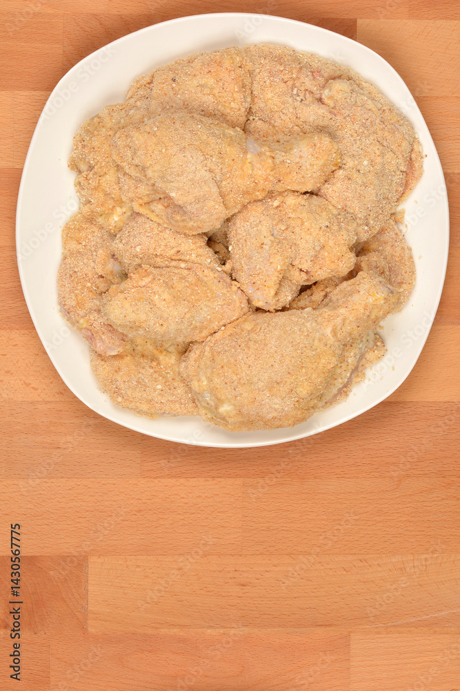 Several pieces of breaded raw chicken, including drumsticks and cutlets, are arranged on a white plate ready for cooking