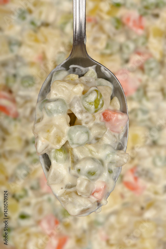 A spoonful of creamy Croatian-style French salad with diced vegetables and mayonnaise is held over the remaining salad in a blurred background