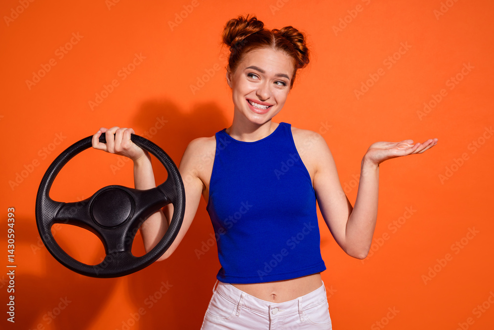 Confident young woman with stylish hairstyle holding a car steering wheel while gesturing, smiling playfully, on an orange background.