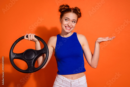 Confident young woman with stylish hairstyle holding a car steering wheel while gesturing, smiling playfully, on an orange background.