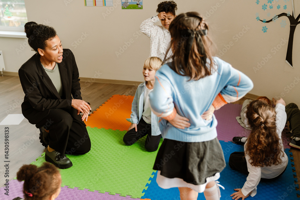 A multiracial group of young children and a female teacher interact on colorful foam mats during a primary school classroom activity.
