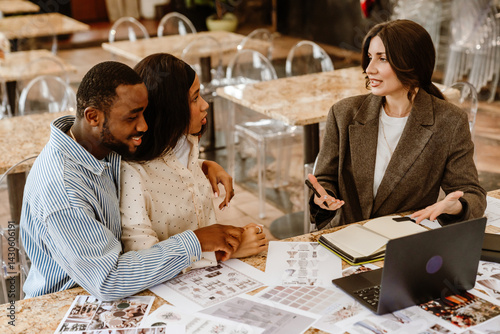 A black adult couple is sitting with a white female wedding planner, discussing ideas and reviewing documents on a laptop and paper in a modern indoor space.